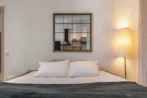 Un lit avec une literie blanche, une lampe de chevet et une fenêtre avec des stores.