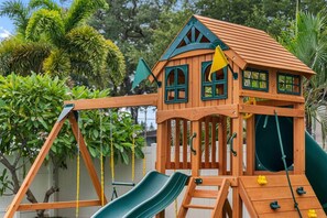A wooden playhouse with a slide, climbing net, and a small tower with windows. - Playcation Oasis (Pinellas County)