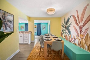 Un coin repas avec une table dressée pour quatre personnes, une cuisine en arrière-plan et une grande fresque murale représentant des motifs végétaux.