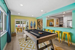 Une table de billard avec des billes, des tabourets jaunes, un coin cuisine et un espace repas avec des chaises.