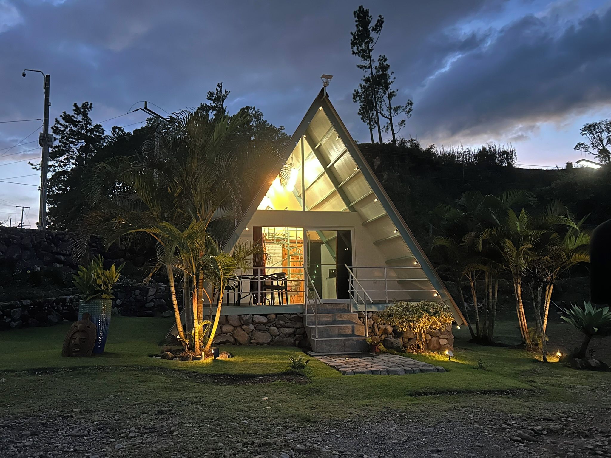 A modern, triangular-shaped building with a glass facade, surrounded by tropical vegetation and a stone wall.