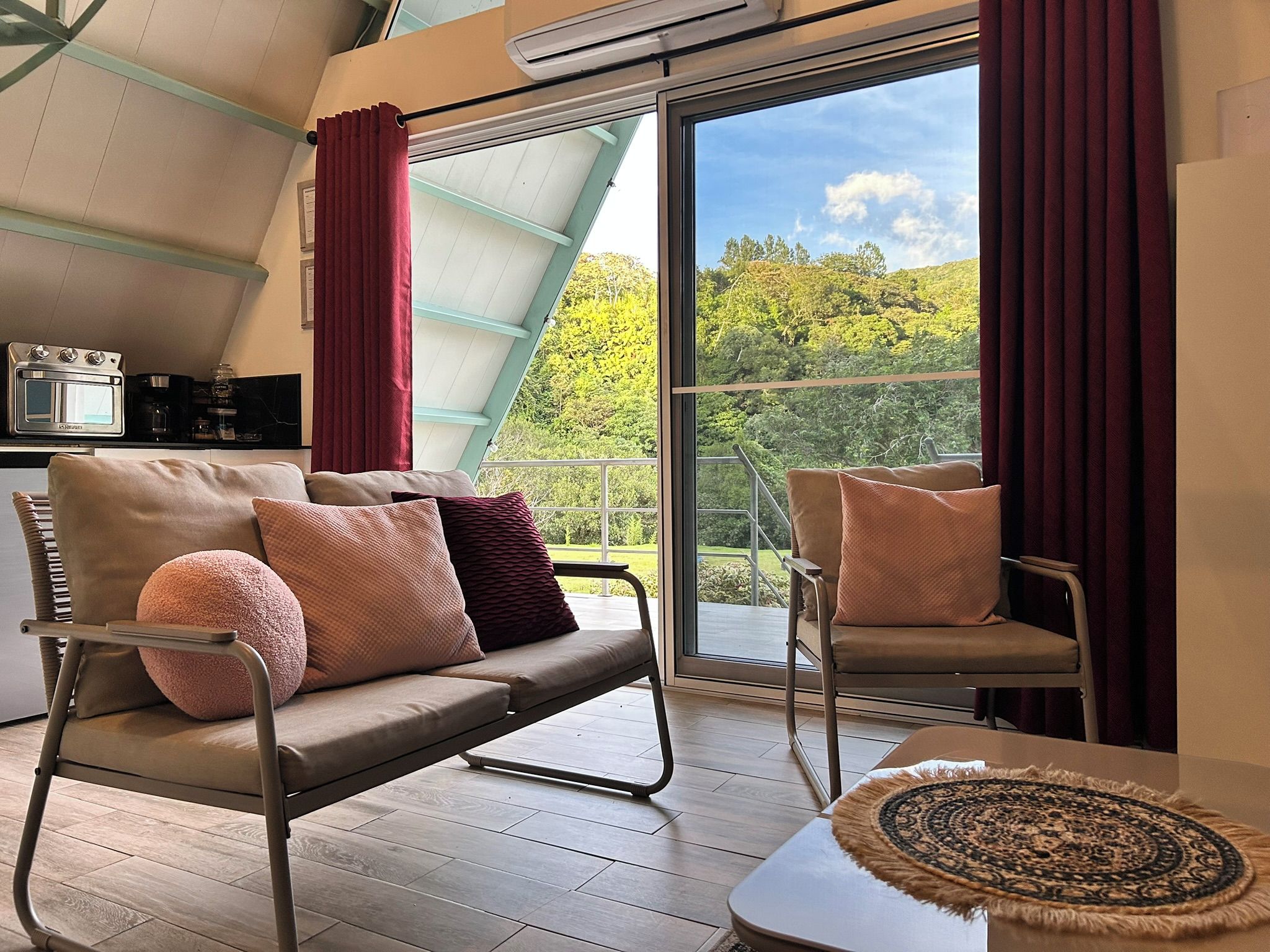Un salon moderne avec un canapé, des fauteuils et une petite table. Il y a une grande fenêtre avec des rideaux, un ventilateur de plafond et un coin cuisine équipé d’appareils électroménagers.