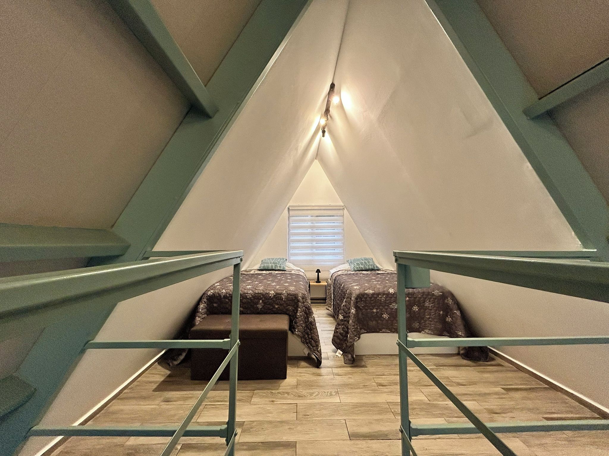 A triangular attic bedroom with two beds, a window with blinds, and a wooden floor.