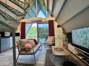 A modern living room with a sofa, chairs, a TV, and a view of greenery through large windows.