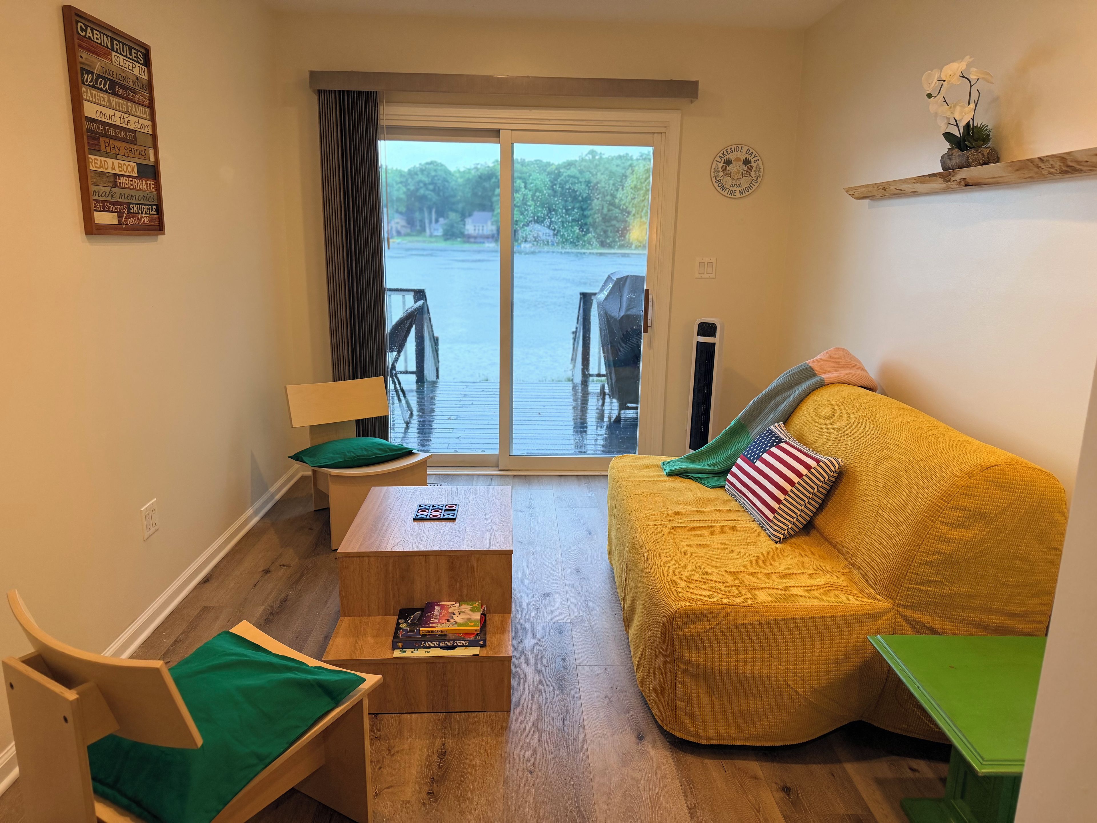 A cozy living room with a yellow sofa, a wooden chair, a small table with books, and a view of a dock through a glass door.