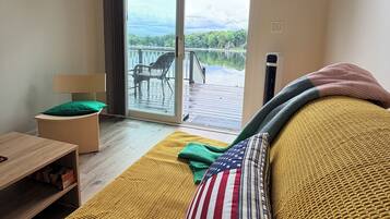 A room with a sofa, a table, a chair, and a view of a lake through a sliding glass door.