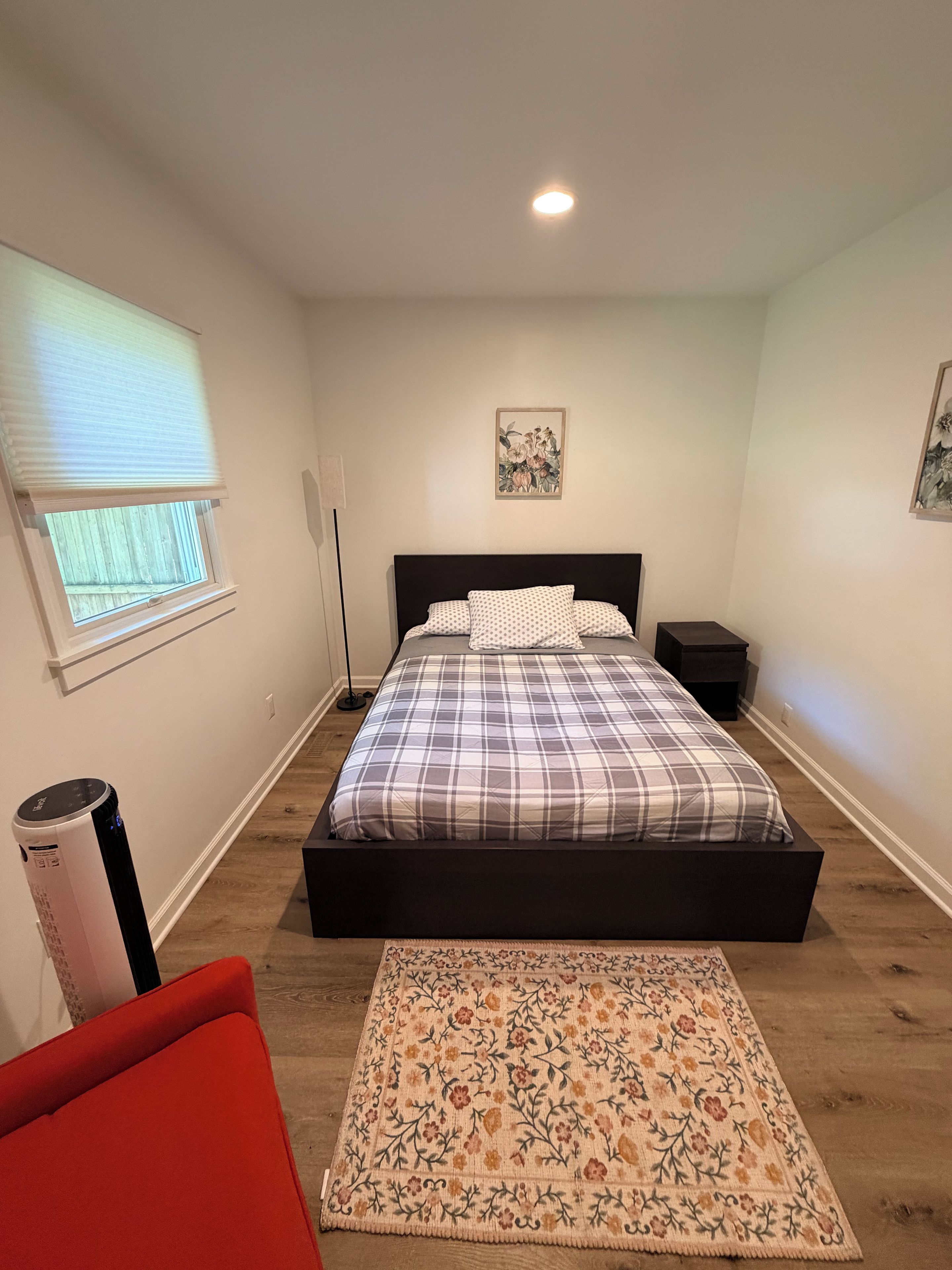 A bedroom with a bed, a bedside table, a nightstand, a window with blinds, a floor lamp, a rug, and a red chair.