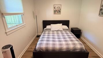 A bedroom with a bed, a bedside table, a nightstand, a window with blinds, a floor lamp, a rug, and a red chair.
