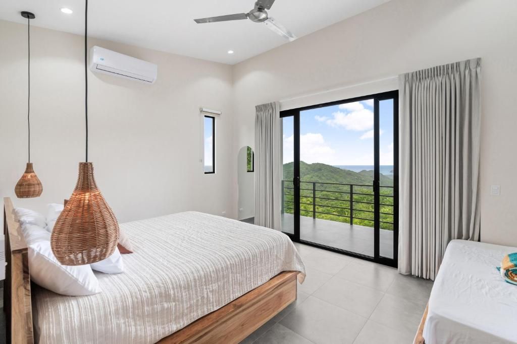 Un dormitorio con una cama grande, un ventilador de techo, un equipo de aire acondicionado y vistas a un paisaje verde a través de una puerta corrediza de vidrio.