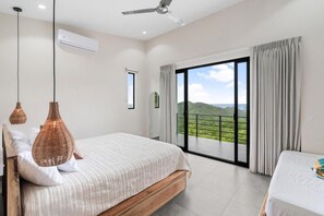 A bedroom with a large bed, a ceiling fan, an air conditioning unit, and a view of a green landscape through a sliding glass door. - Villas Vivo (Potrero)