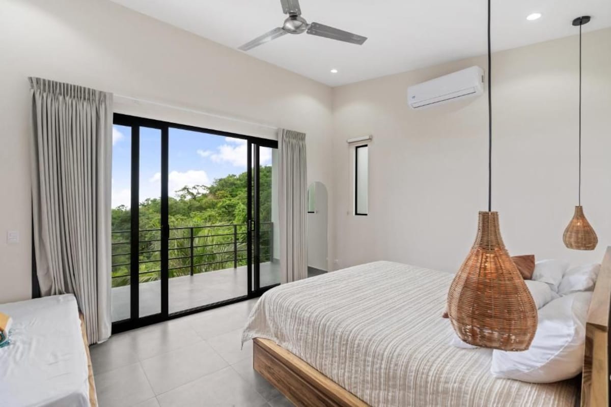 Un dormitorio moderno con una cama grande, ventilador de techo y vistas a zonas verdes a través de puertas correderas de cristal.