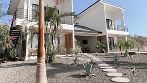 Una casa moderna de dos pisos con un balcón, rodeada por un camino de grava y plantas tropicales.