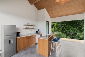 Una cocina moderna con refrigerador empotrado, cafetera y microondas. Hay una barra de bar de madera con taburetes y un ventanal con vistas a la vegetación.