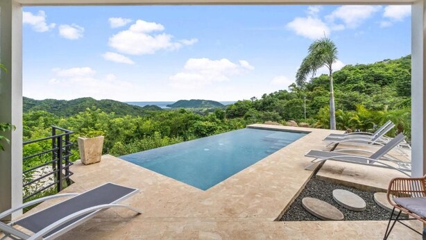 Una piscina infinita rectangular con sillas de descanso y un camino de piedra, rodeada de exuberante vegetación y colinas.