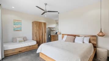 Un dormitorio con una cama grande, una cama más pequeña, un armario, un ventilador de techo y una pequeña kitchenette.