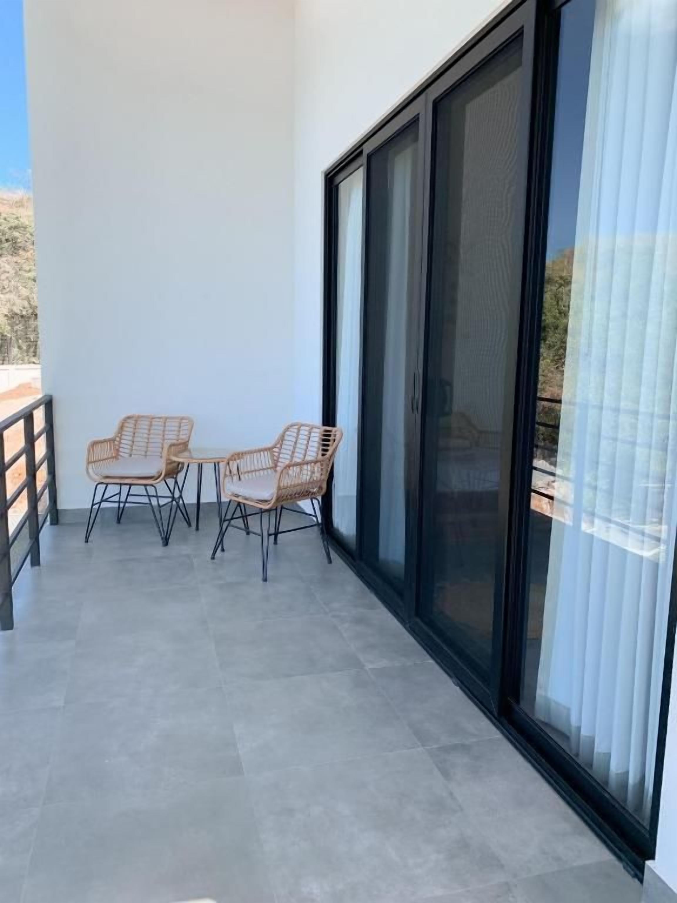 A balcony with two wicker chairs and a table, a sliding glass door, and a tiled floor.