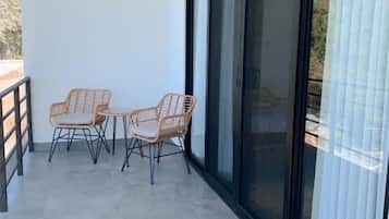 A balcony with two wicker chairs and a table, a sliding glass door, and a tiled floor.