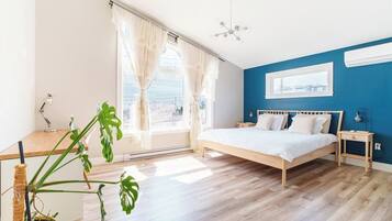 Une chambre à coucher avec un lit, des tables de chevet, une fenêtre avec des rideaux, une plante en pot et une vue sur l’extérieur.