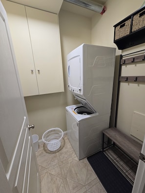 A laundry room with a front-loading washing machine, a drying rack, a small bench, and a white basket.