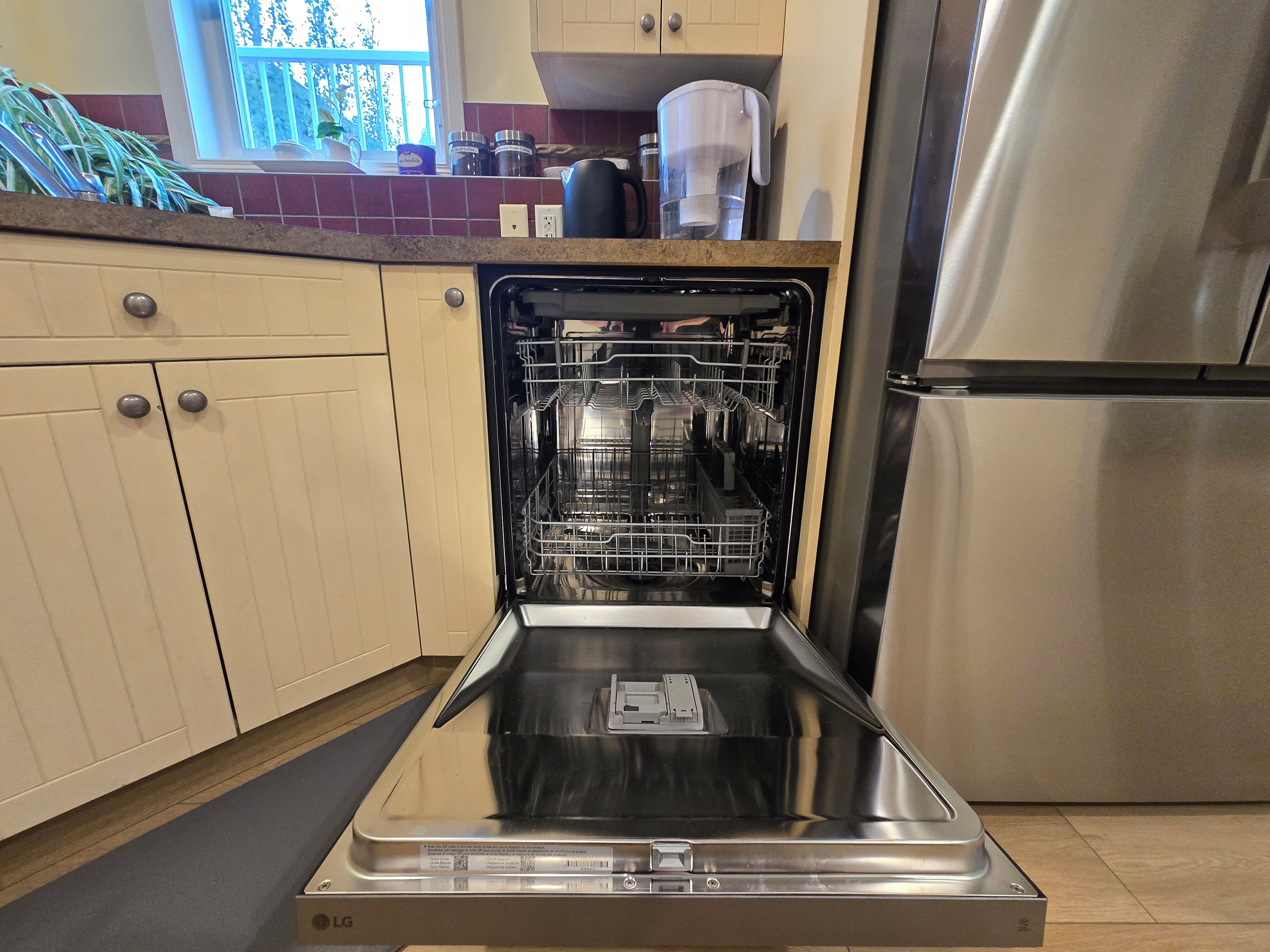 Un lave-vaisselle ouvert dans une cuisine équipée d’un réfrigérateur en acier inoxydable et de meubles de couleur beige.