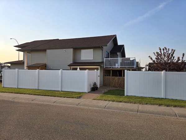 A two-story residential house with a front deck, a white picket fence, and a clear sky.