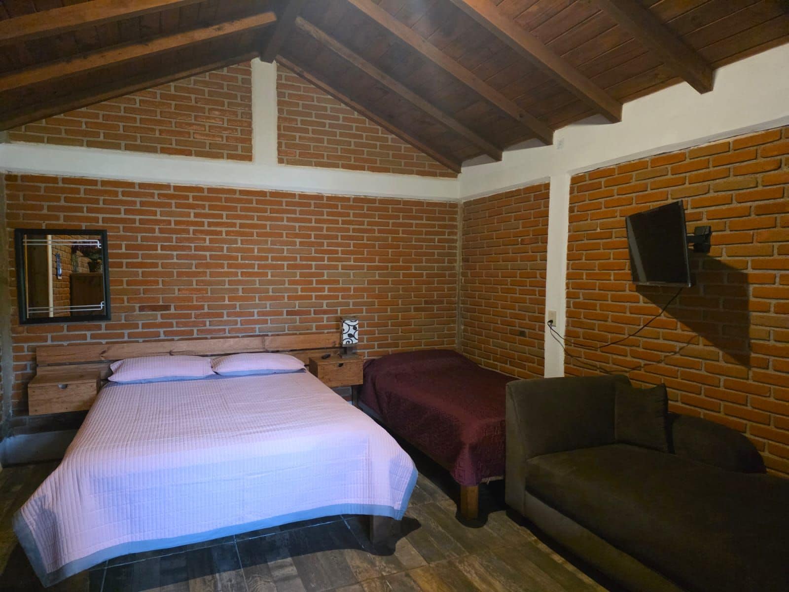 Ein Zimmer mit Backsteinwand, einem Bett mit weißer Bettdecke, einem hölzernen Kopfteil, einem Nachttisch, einem an der Wand montierten Fernseher und einem braunen Sofa.
