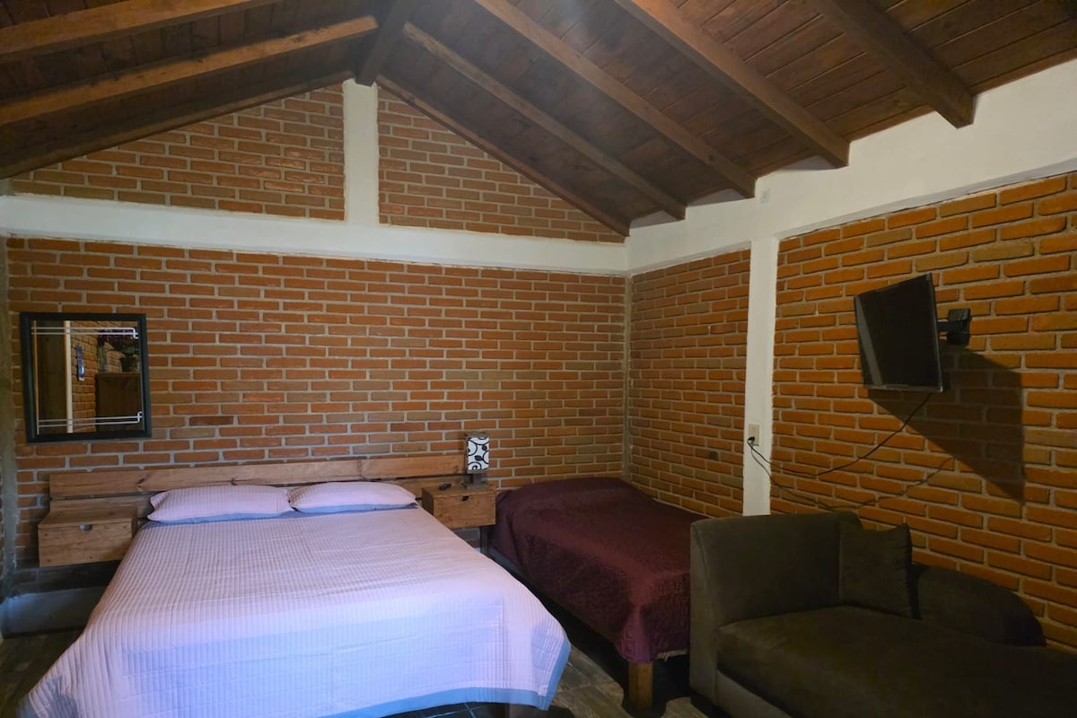 Una habitación con pared de ladrillo, una cama con colcha blanca, cabecero de madera, una mesita de noche, un televisor montado en la pared y un sofá marrón.