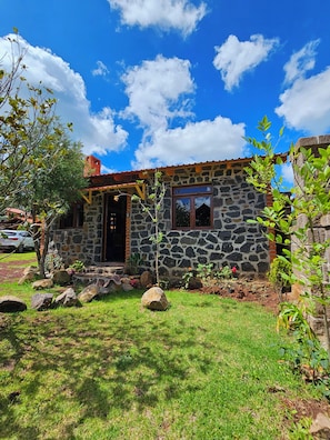 A stone house with a red roof, a wooden door, and a small garden with rocks and plants. - Cabañas JH (Huasca)
