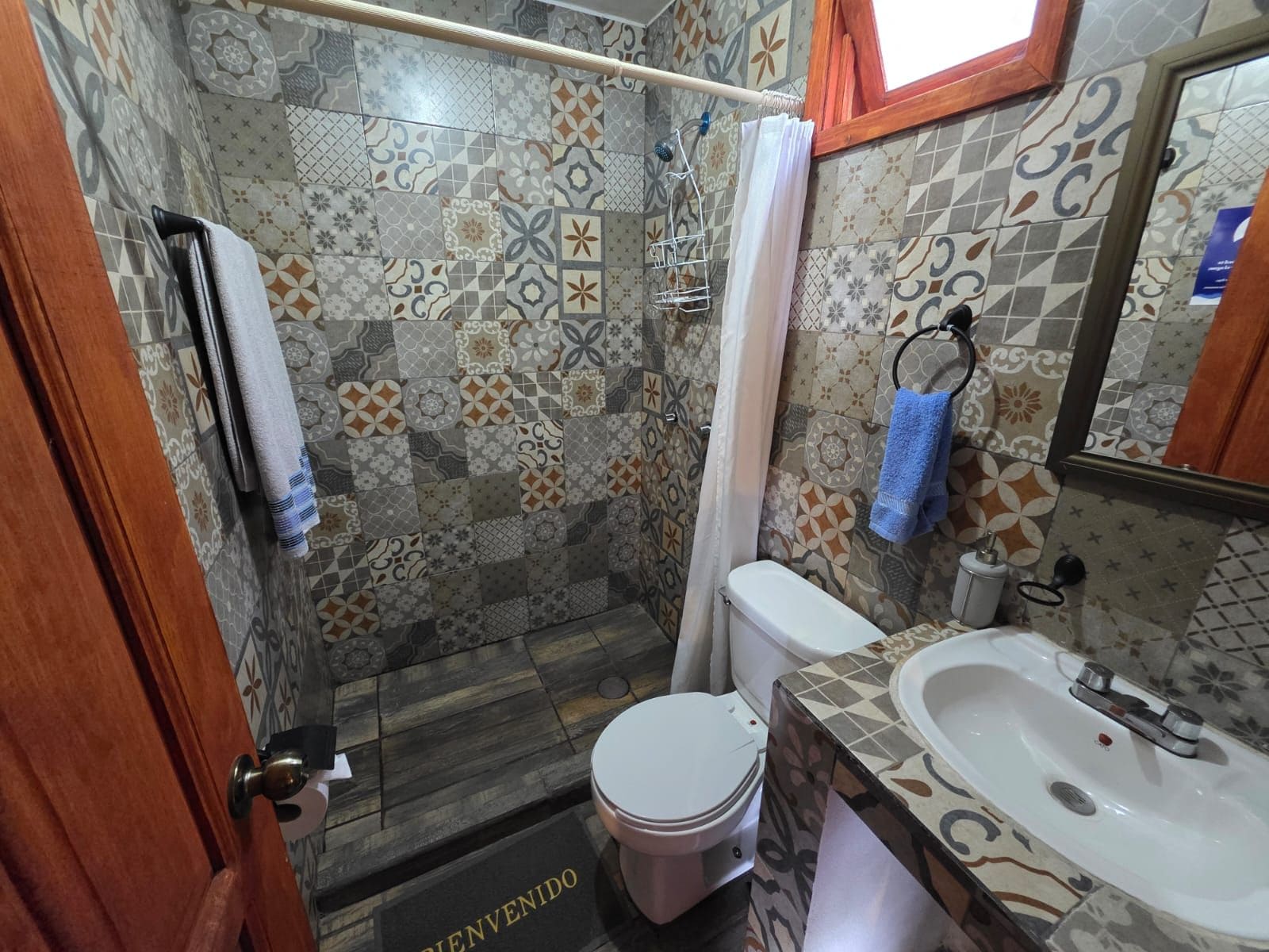 Un baño con ducha, inodoro y lavamanos, con azulejos estampados y una puerta de madera.