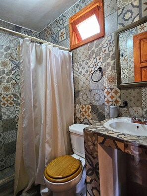 Un baño con cortina de ducha, inodoro, lavamanos y una ventana sobre el espejo.