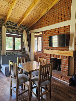 A cozy room with a brick fireplace, a television mounted above it, a dining table with chairs, and a stone wall. - Cabañas JH (Huasca)