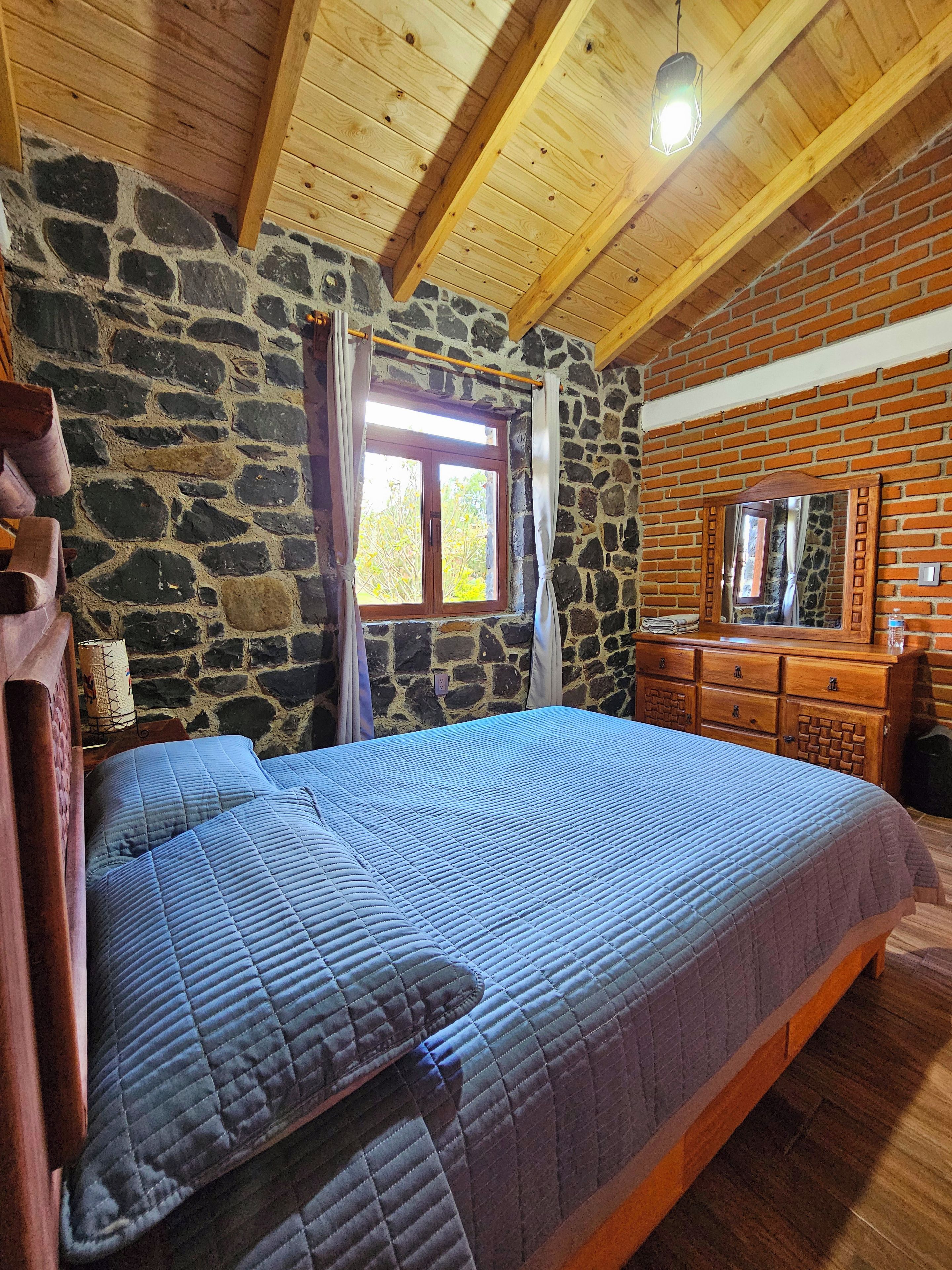 A bedroom with a stone wall, a wooden ceiling, a bed with a blue quilt, a wooden dresser, and a window with curtains.