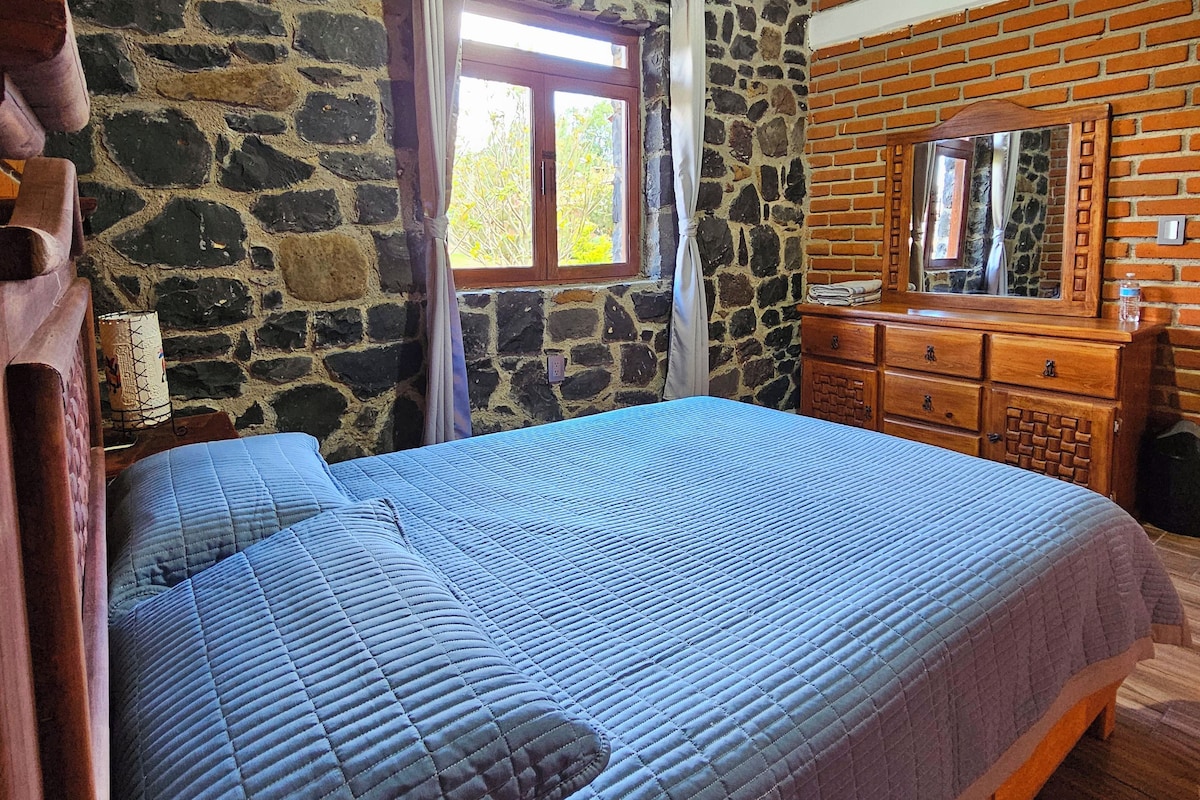 Un dormitorio con pared de piedra, techo de madera, cama con colcha azul, cómoda de madera y ventana con cortinas.