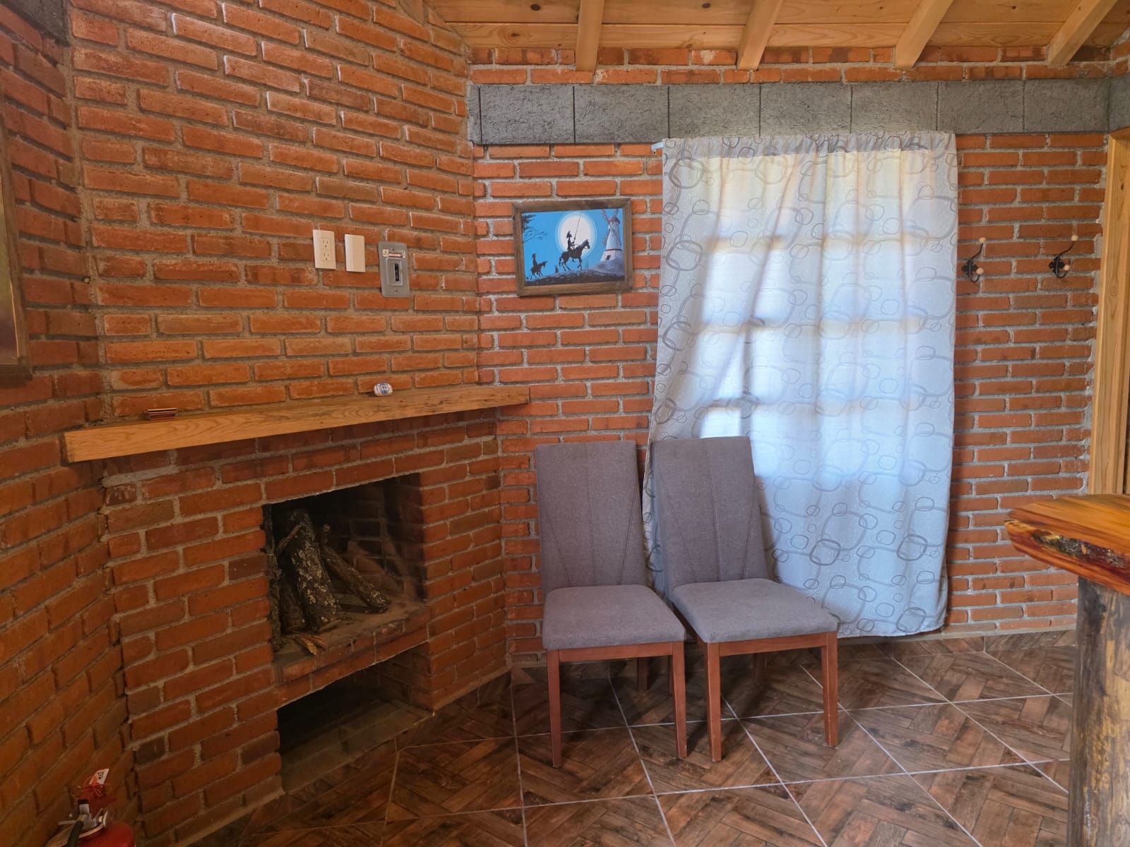 A room with a brick fireplace, two chairs, a wooden shelf, and a patterned curtain.