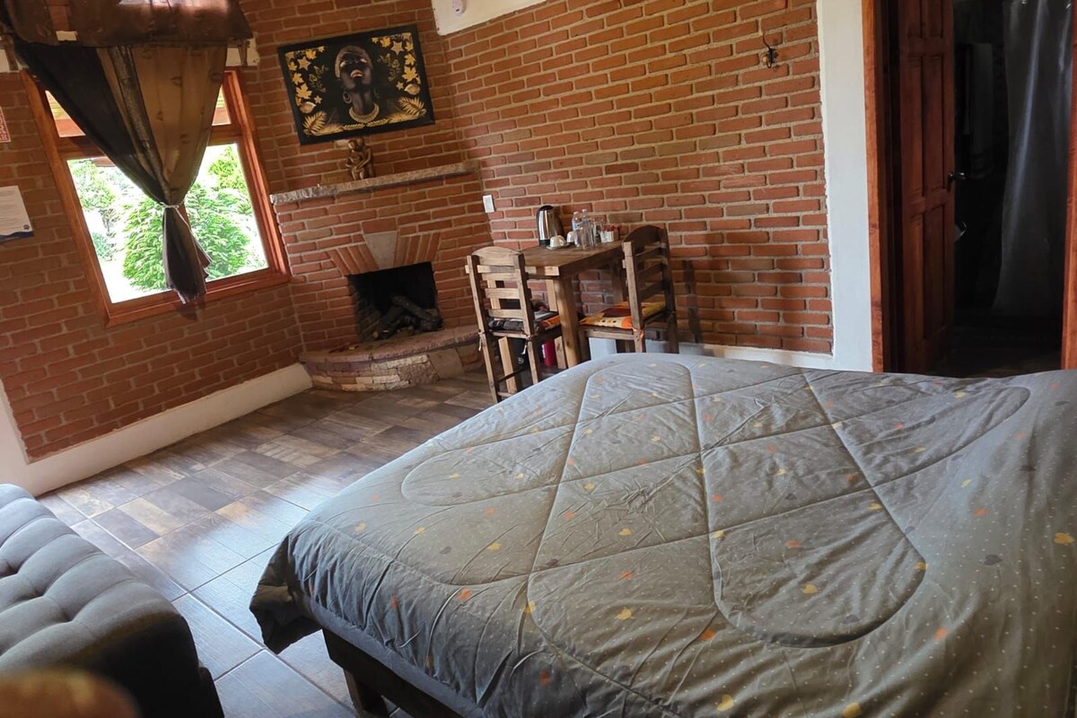 Una habitación con chimenea de ladrillo, una cama con colcha, una mesa con sillas y un cuadro en la pared.