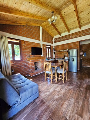 Amplia sala de estar con sofá, mesa de comedor y sillas, chimenea, televisor montado en una pared de ladrillo y refrigerador.