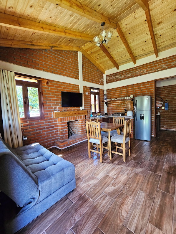 A spacious living area with a sofa, dining table, and chairs, a fireplace, a television mounted on a brick wall, and a refrigerator. - Cabañas JH (Huasca)