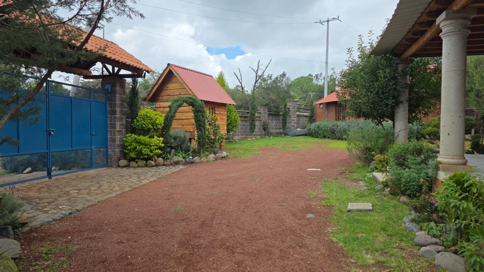 A rustic wooden cabin with a red roof, surrounded by a gravel path and greenery.