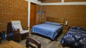 Una habitación con pared de ladrillo, puerta de madera, cama con colcha azul, silla de madera y una mesita.