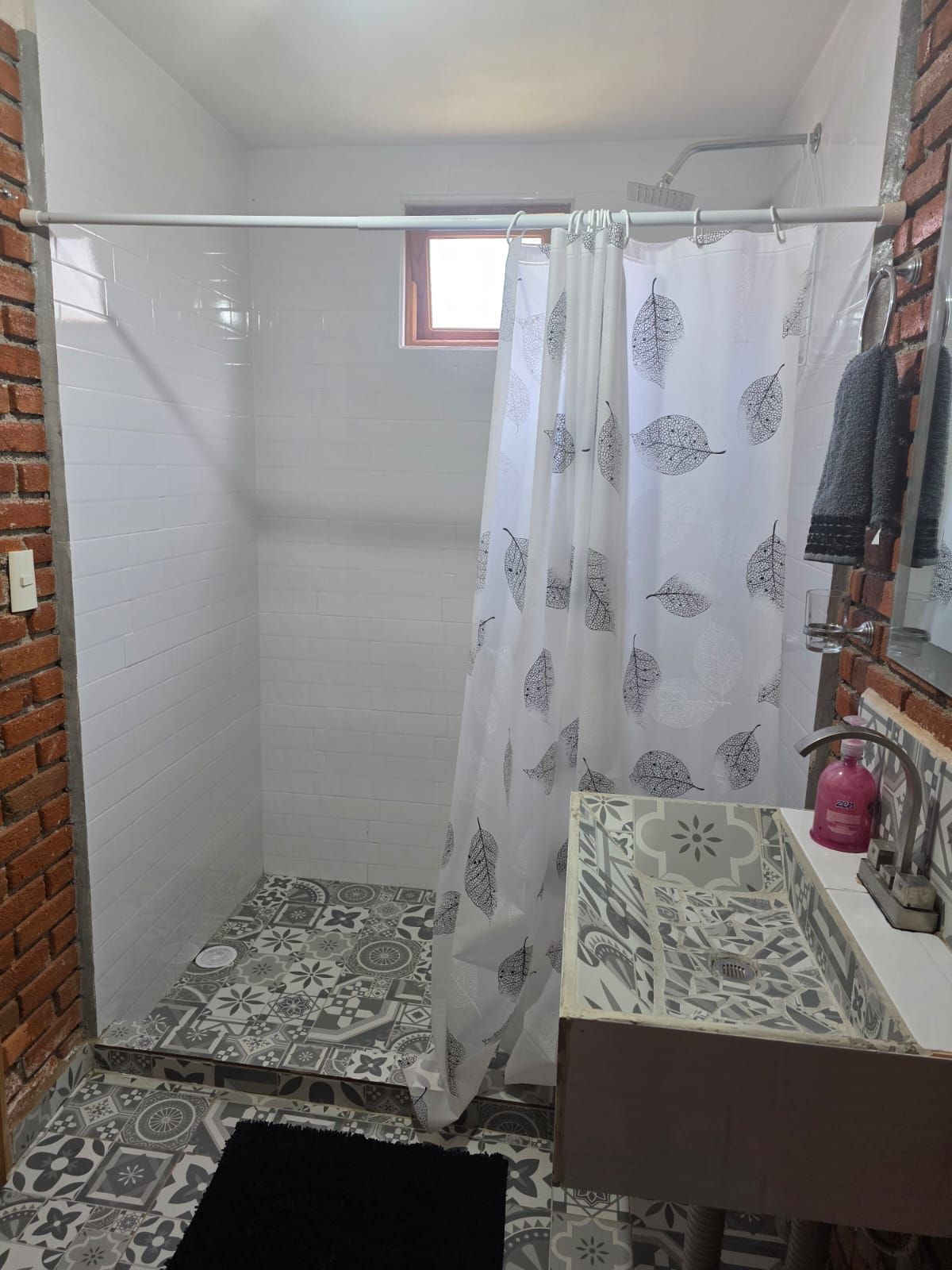A bathroom with a shower, a sink with a patterned tile backsplash, and a window with a curtain.