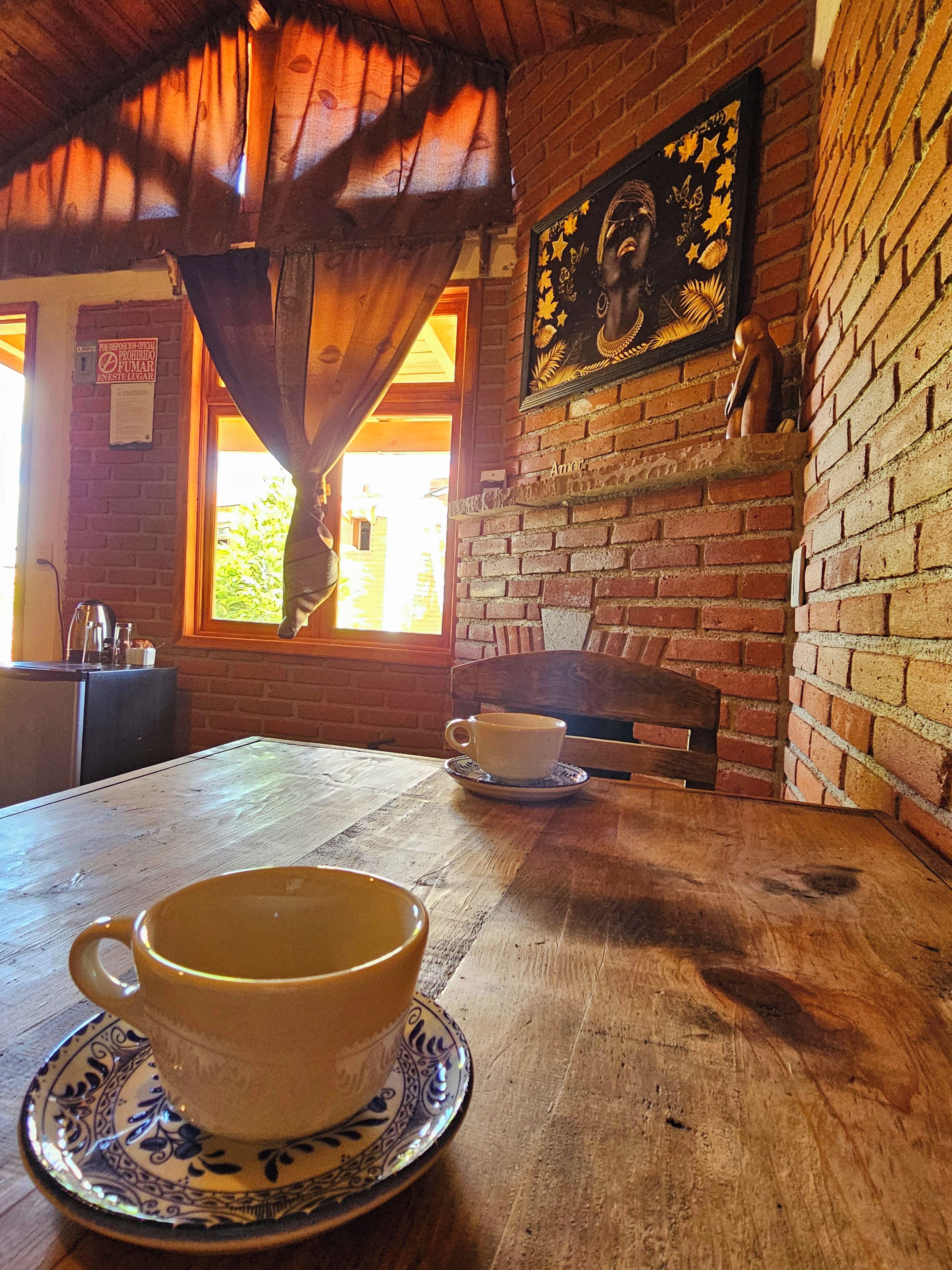 Interior rústico com mesa de madeira, uma xícara em pires, um banco e parede de tijolos com um quadro emoldurado.