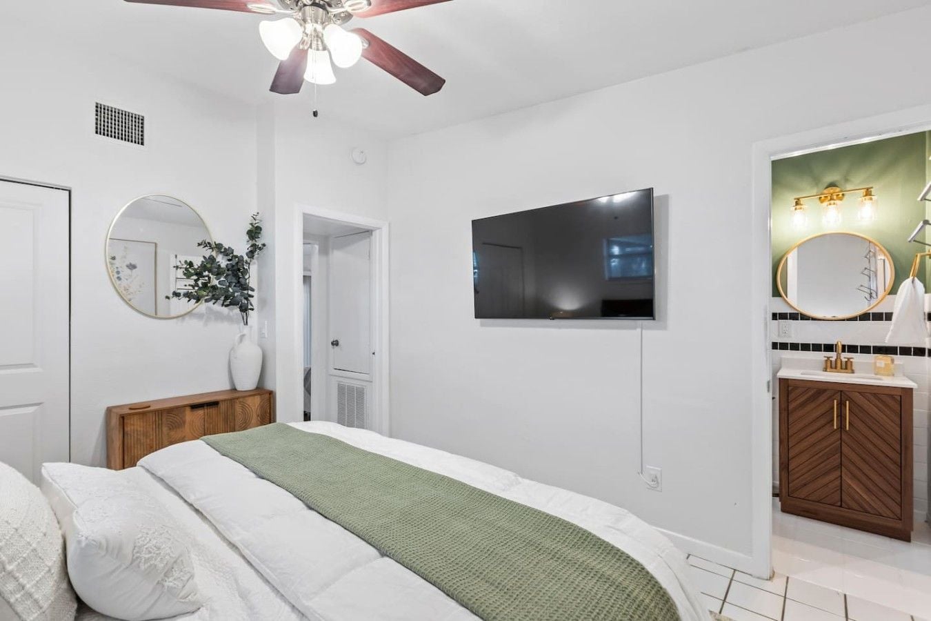 Une chambre à coucher comprenant un lit, un ventilateur de plafond, une télévision, un miroir et une salle de bain avec un lavabo et un meuble-lavabo.