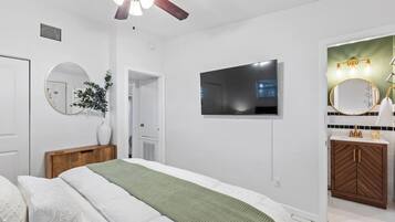Une chambre à coucher comprenant un lit, un ventilateur de plafond, une télévision, un miroir et une salle de bain avec un lavabo et un meuble-lavabo.