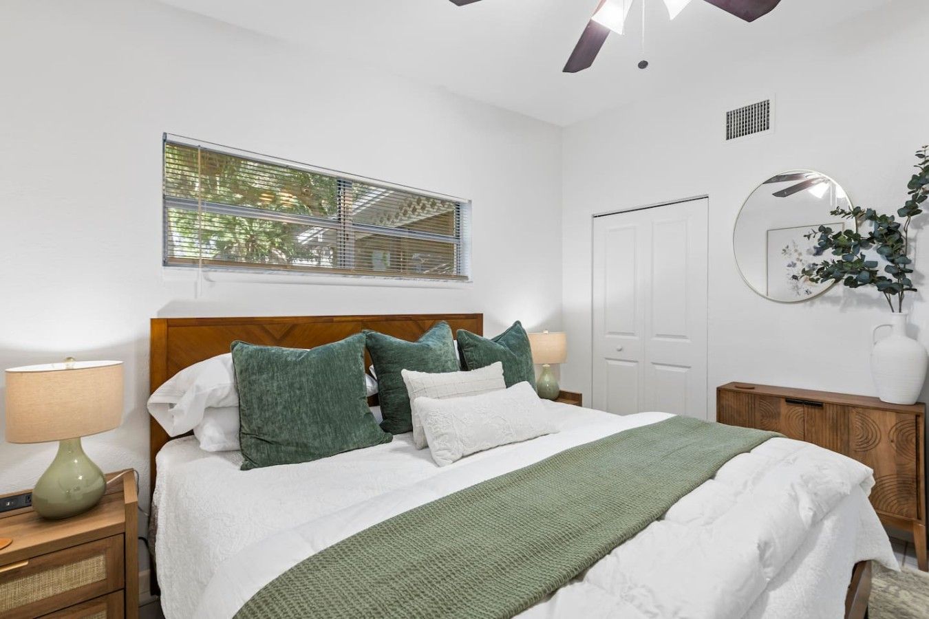 A neatly arranged bedroom with a bed, bedside lamps, a window with blinds, a mirror, and a potted plant.