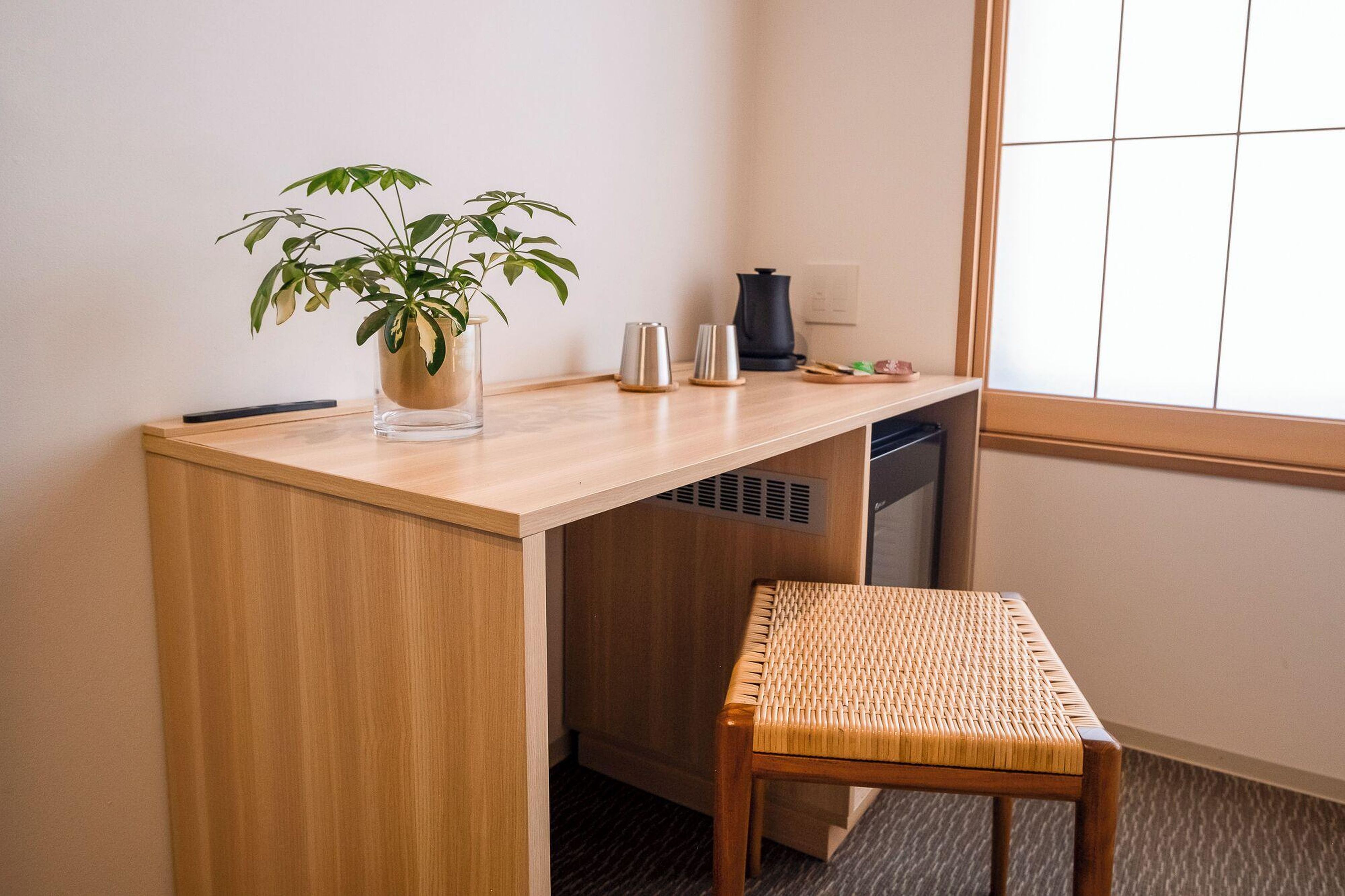 A compact workspace with a wooden desk, a clear glass vase with a plant, a small metal kettle, and a woven chair.
