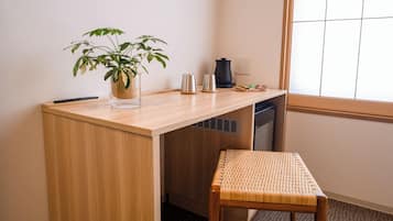 A compact workspace with a wooden desk, a clear glass vase with a plant, a small metal kettle, and a woven chair.