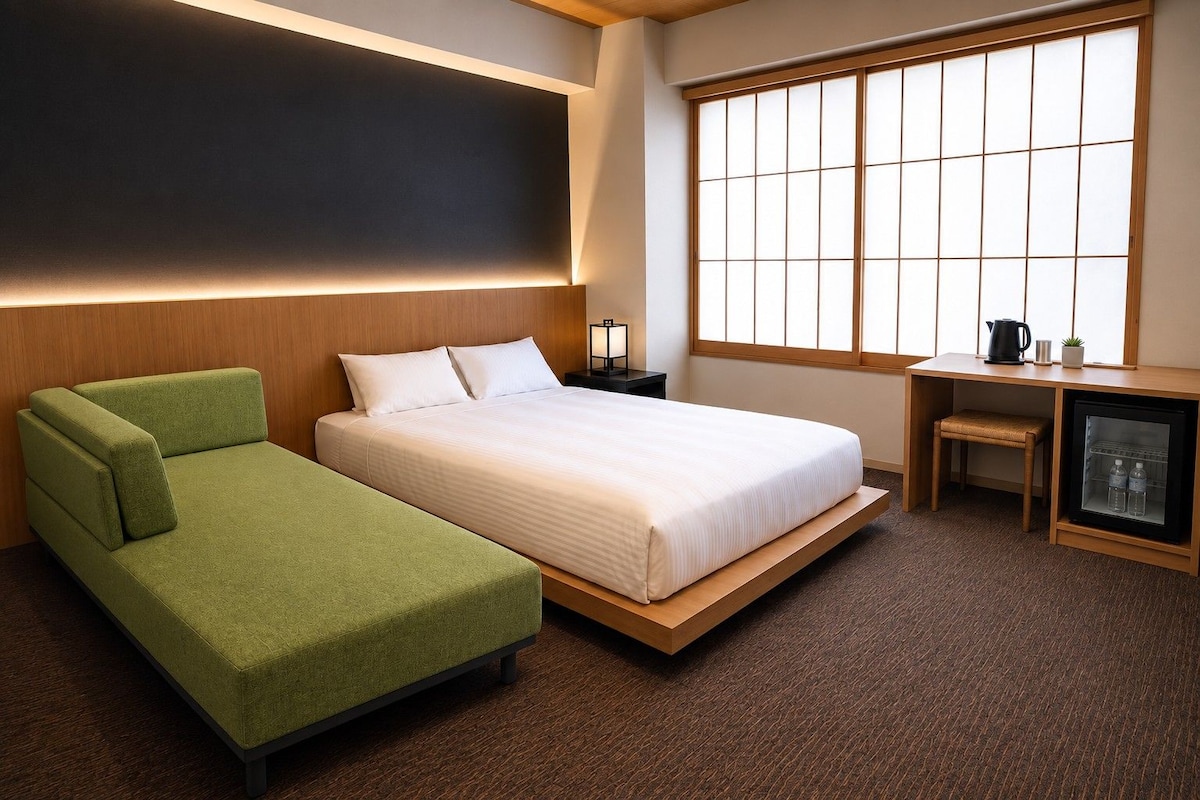 A hotel room with a bed, a green sofa, a small table, a cabinet, and a large window.