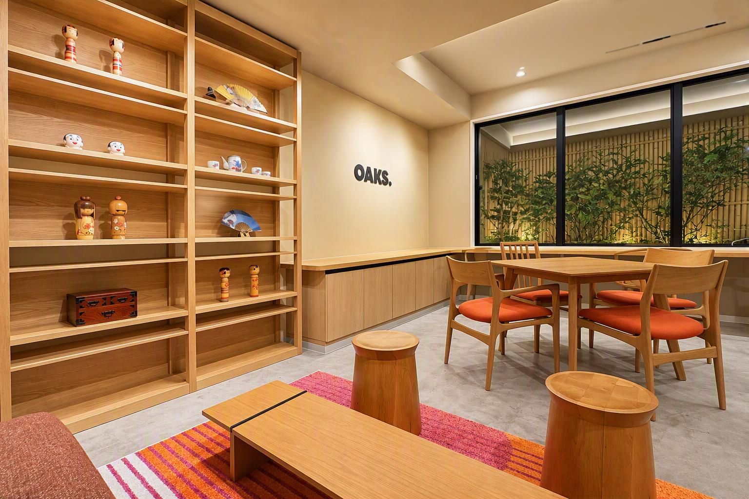 Un interior moderno con una estantería de madera, un comedor con sillas anaranjadas y un ventanal con vistas al verdor.