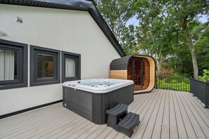 Ein moderner Whirlpool auf einer Holzterrasse, eine Holzsagastruktur und ein weißes Gebäude mit schwarzen Fensterrahmen.