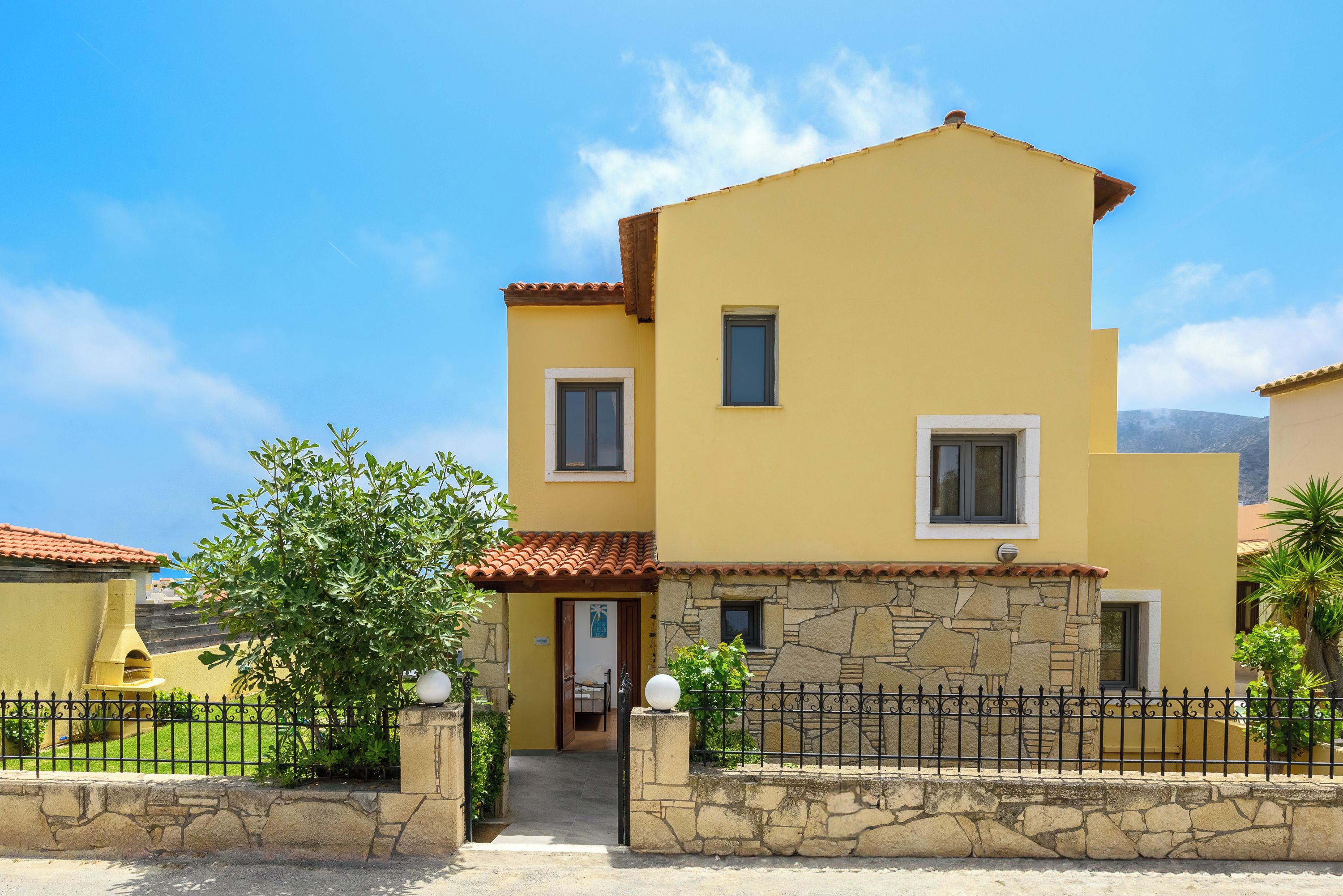 A two-story yellow building with a stone facade, a tiled roof, and a black metal fence.